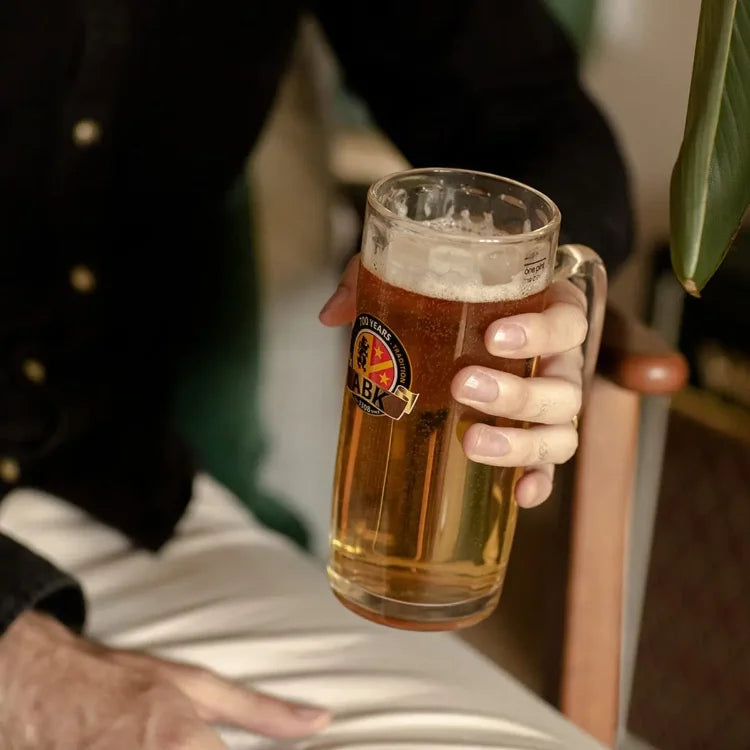 A man holds a pint of abk hell beer while sat down